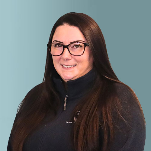 A person with long dark hair and glasses, wearing a dark zip-up jacket, stands against a solid light teal background. They are smiling gently and looking at the camera.