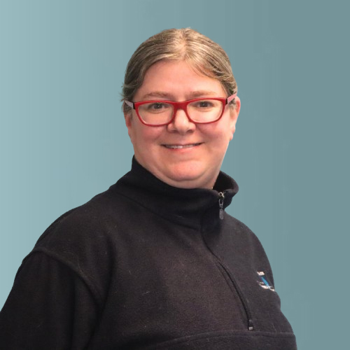 A person with short hair and glasses is smiling, wearing a black zip-up jacket against a plain blue background.