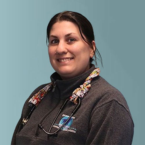 A person with dark hair is smiling and wearing a dark gray zip-up jacket with a colorful lanyard holding a stethoscope around their neck. The background is a solid light blue color.