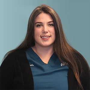 A woman with long brown hair, wearing a blue scrub uniform and a black cardigan, smiles against a light blue background. She has a nose ring and a confident expression.