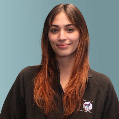 A person with long brown hair smiles softly while wearing a black fleece with embroidered logos. The background is a muted teal color.
