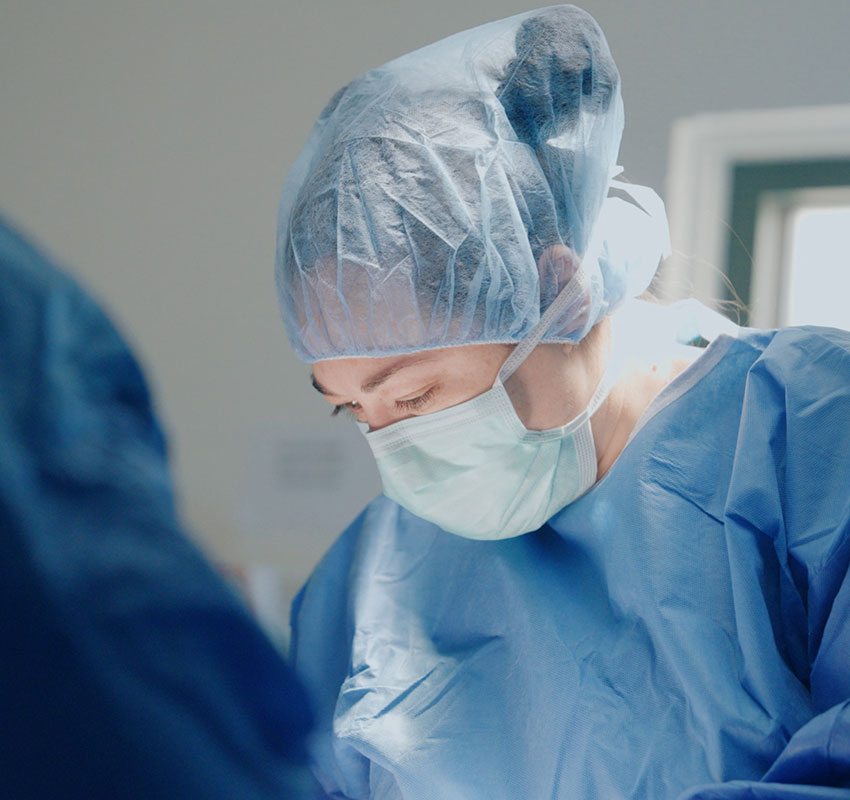 A surgeon wearing a blue surgical gown, cap, and mask is focused on performing a medical procedure in a brightly lit operating room.